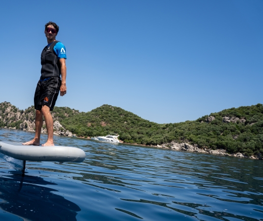 a person standing barefoot on a gray electric hydrofoil surfboard floating on calm, deep blue water.