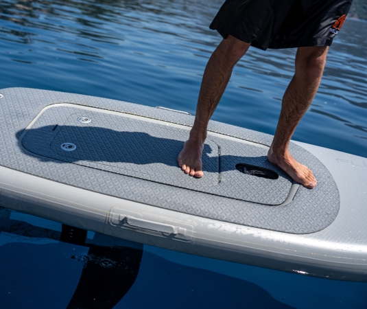 a person standing barefoot on a gray electric hydrofoil surfboard floating on calm, deep blue water.