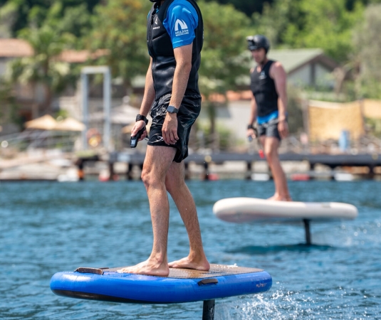 two people riding electric hydrofoil surfboards on a body of water near a shore with greenery and houses. 