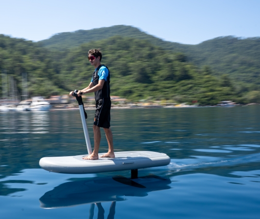 a boy riding a white electric hydrofoil surfboard on calm blue water.