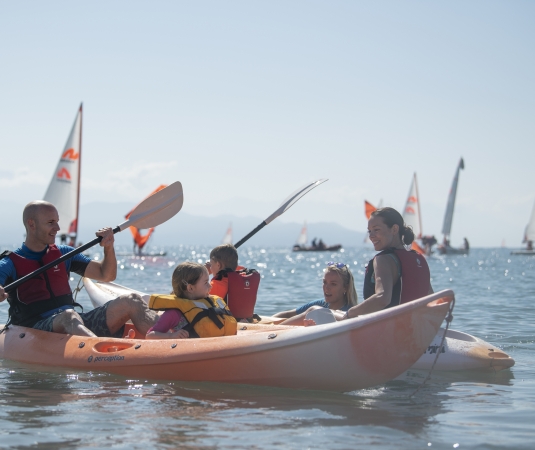 Two adults and three children are seated in a single orange kayak, all wearing life jackets for safety