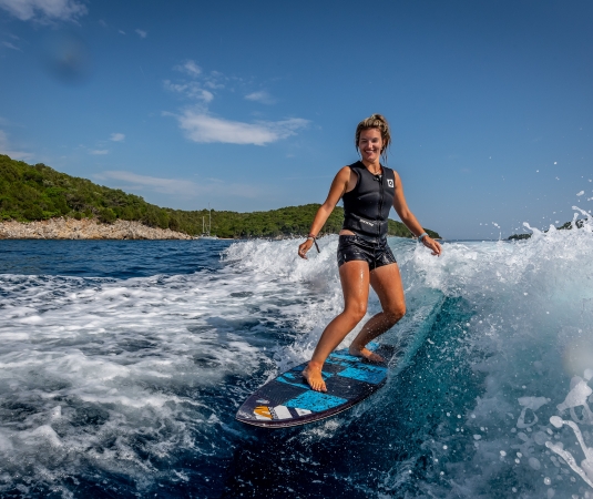 a woman wakesurfing on a blue surfboard in the ocean. She is wearing a black sleeveless top and black shorts, looking confident as she rides the wave.