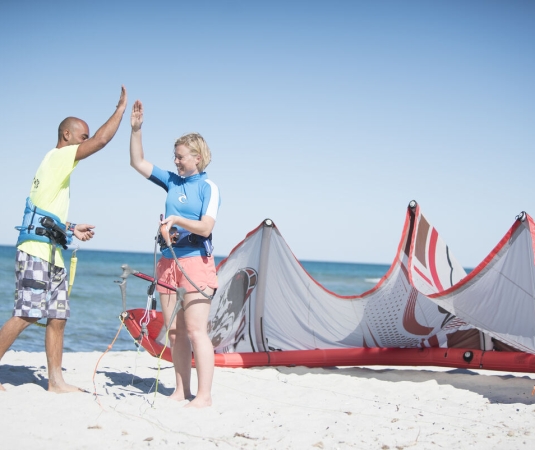 The image shows two people on a sandy beach next to a large, partially deflated red and white kiteboarding kite. A man in a yellow shirt and checkered shorts is giving a high-five to a woman wearing a blue and white top and light pink shorts. Both individuals are barefoot and appear to be preparing for kiteboarding. 