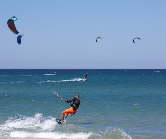 The image shows a vibrant scene of kiteboarding on a sunny, clear day at the sea. In the foreground, a kiteboarder wearing bright orange shorts and a black wetsuit is skillfully riding the waves, holding onto the kite's control bar
