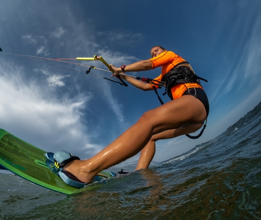 A woman kitesurfing on the ocean, the kite is high in the sky and the photo is taken from a low angle near the water, showing the board clearly