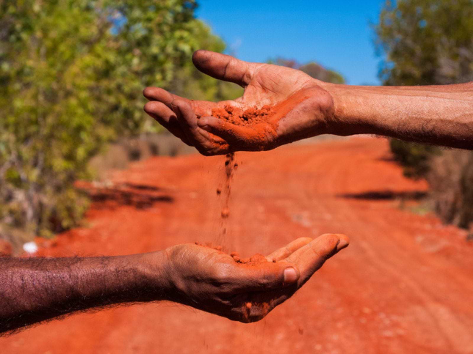 Hånd, der hælder rødt sand ned i anden hånd på rød grusvej i Australien