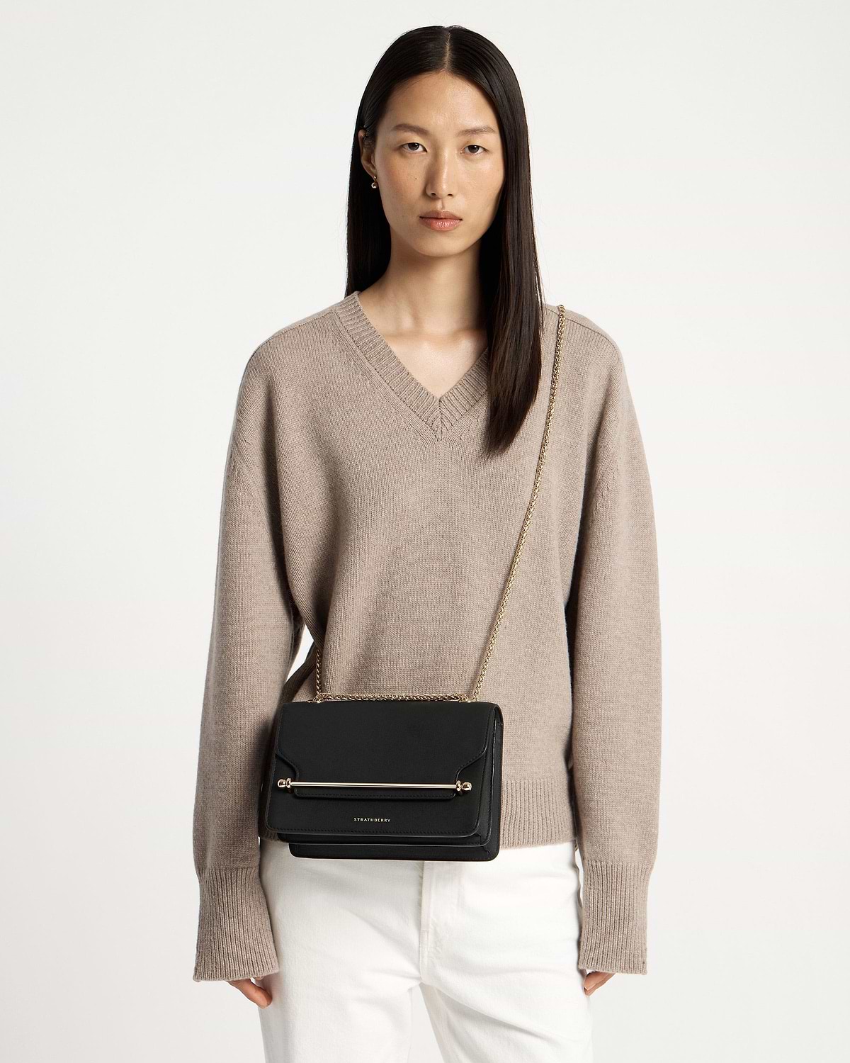 Model wearing black Strathberry east-west crossbody bag with gold bar and chain strap.
