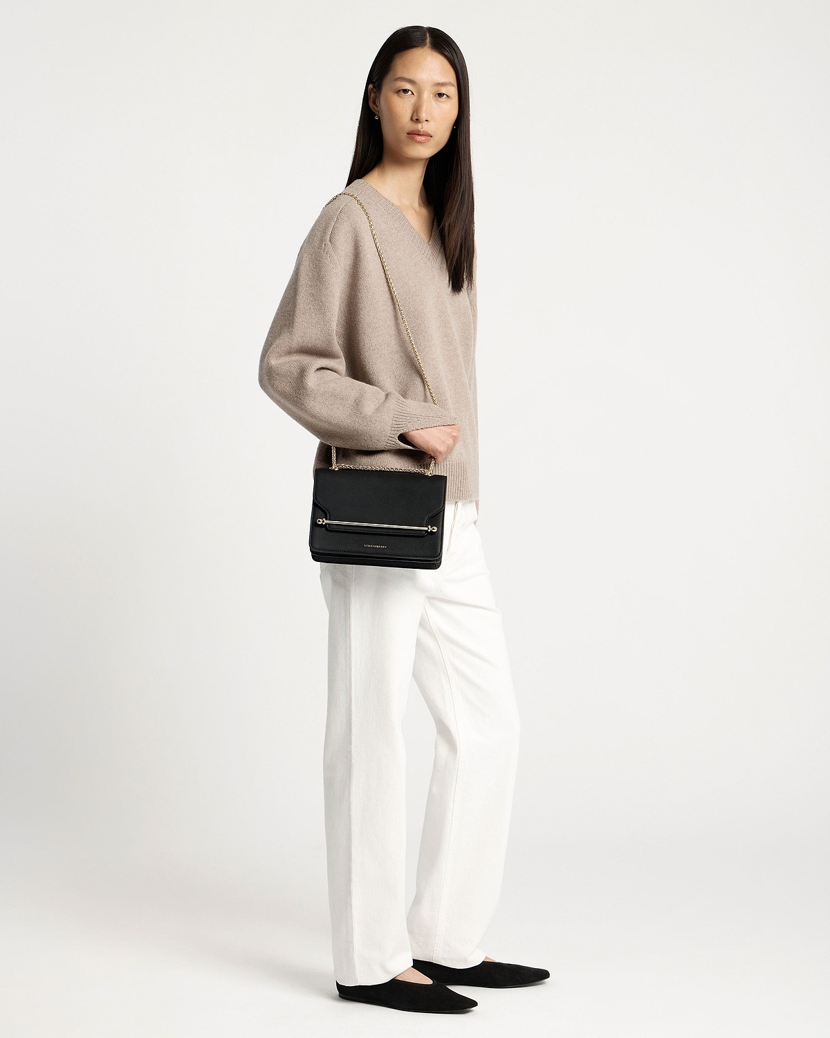 Model wearing black east-west shoulder bag with gold chain and boxy flap.