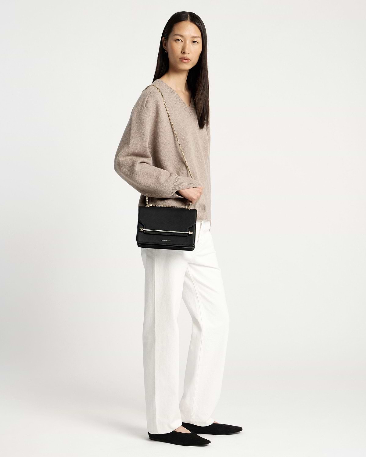 Model wearing black east-west shoulder bag with gold chain and boxy flap.