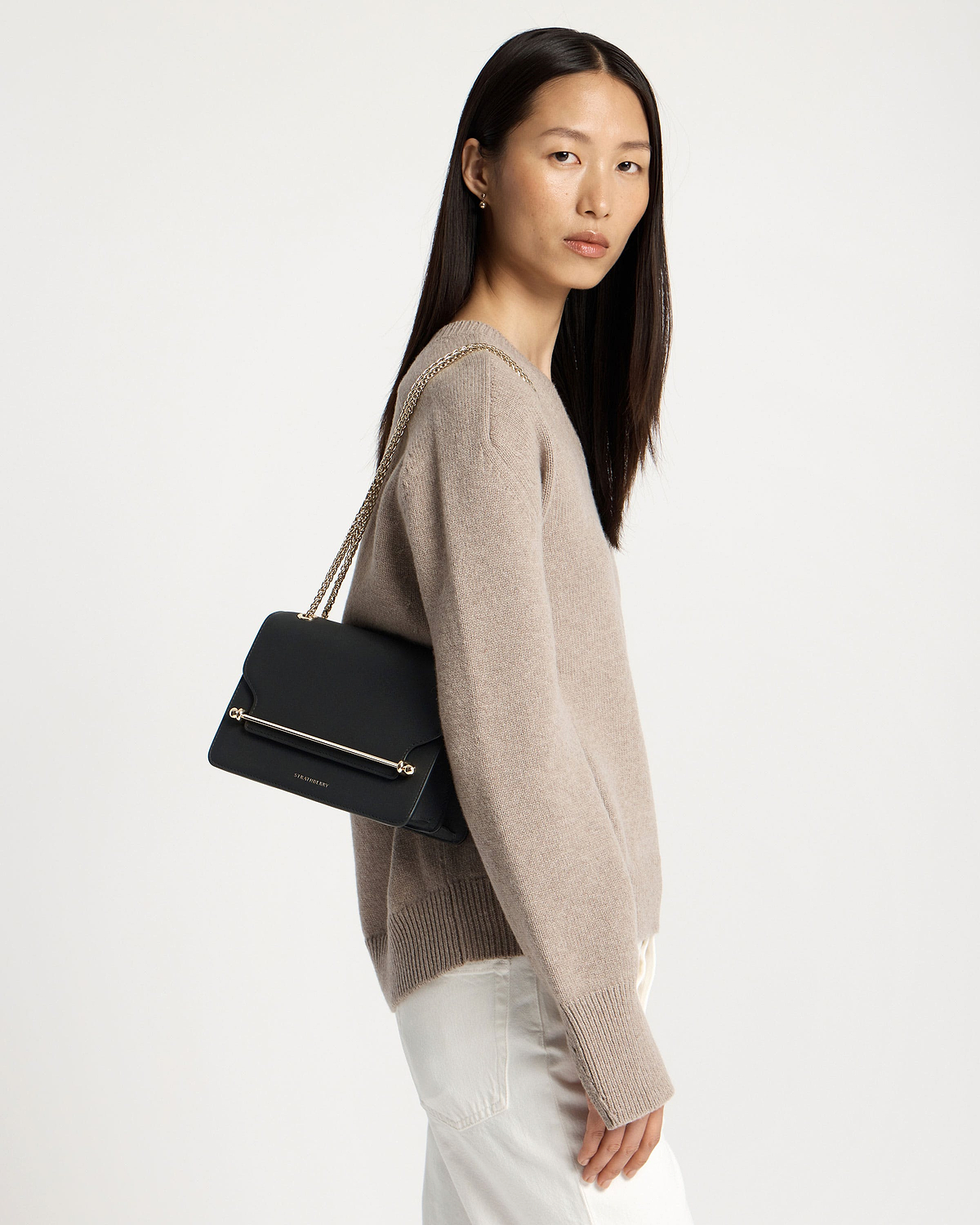 Model wearing beige sweater carrying black east-west bag with gold chain strap and bar.