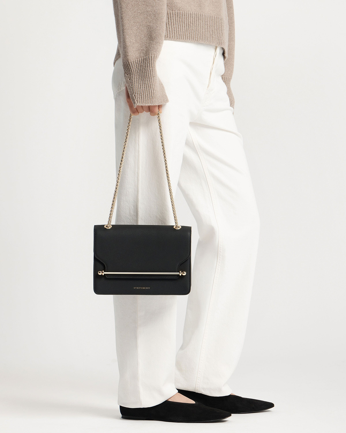 Model holding black Strathberry East/West bag with gold chain strap and bar.