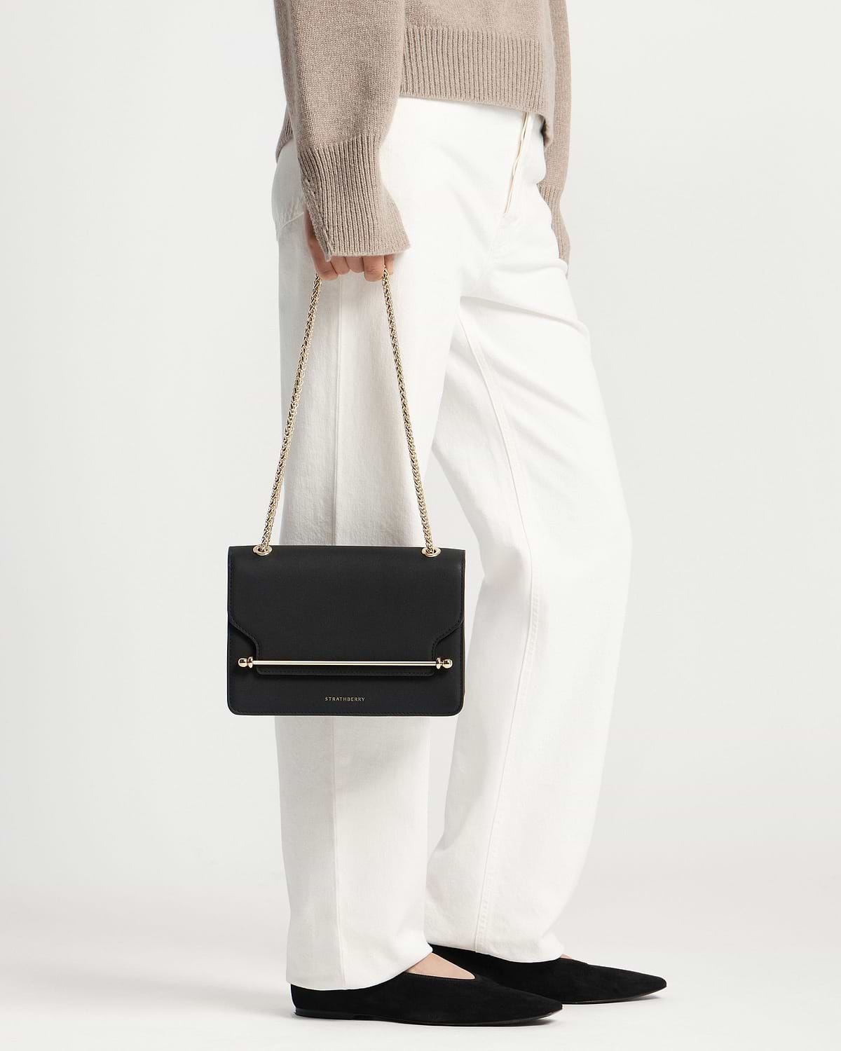 Model holding black Strathberry East/West bag with gold chain strap and bar.
