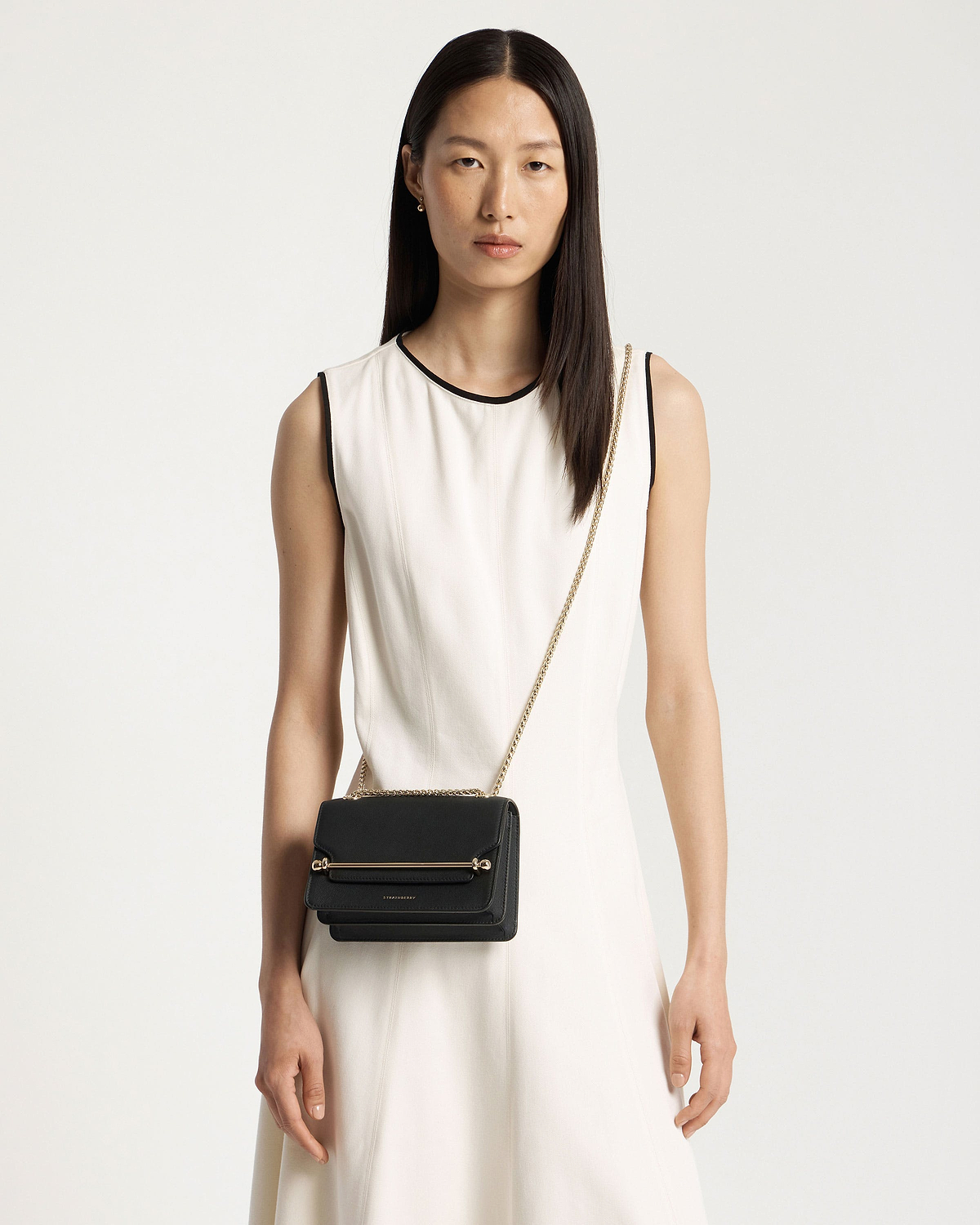 Model in sleeveless white dress wearing black East/West Mini crossbody bag with gold chain