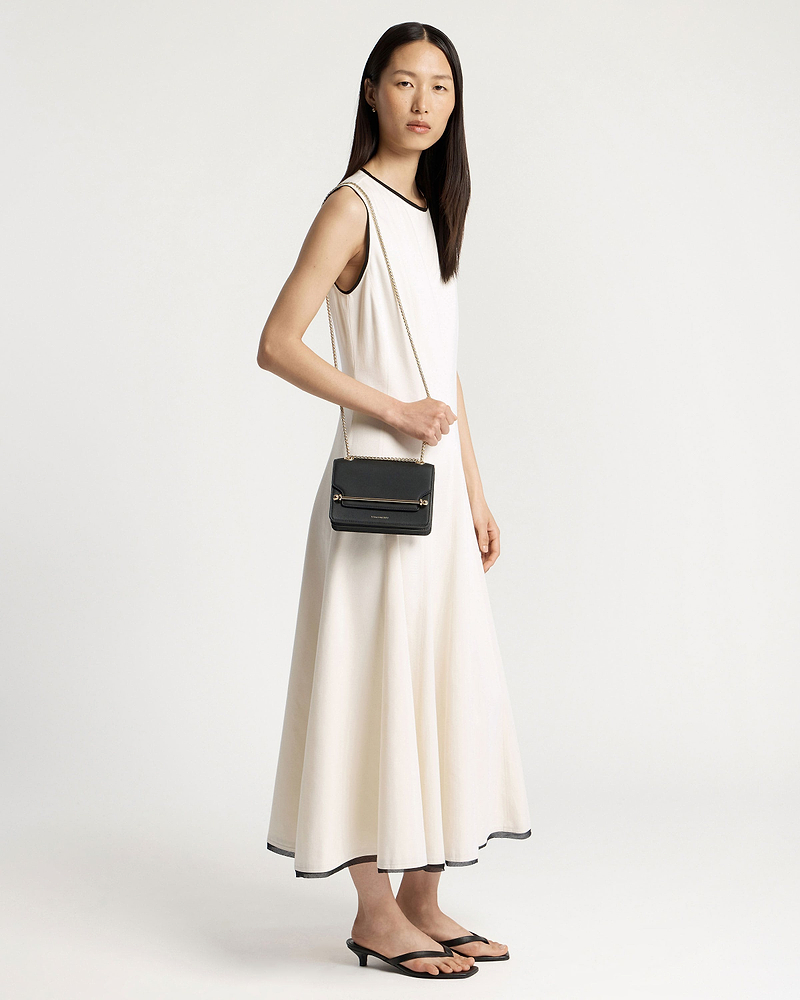 Model in a white sleeveless dress holding a small black crossbody bag with chain.