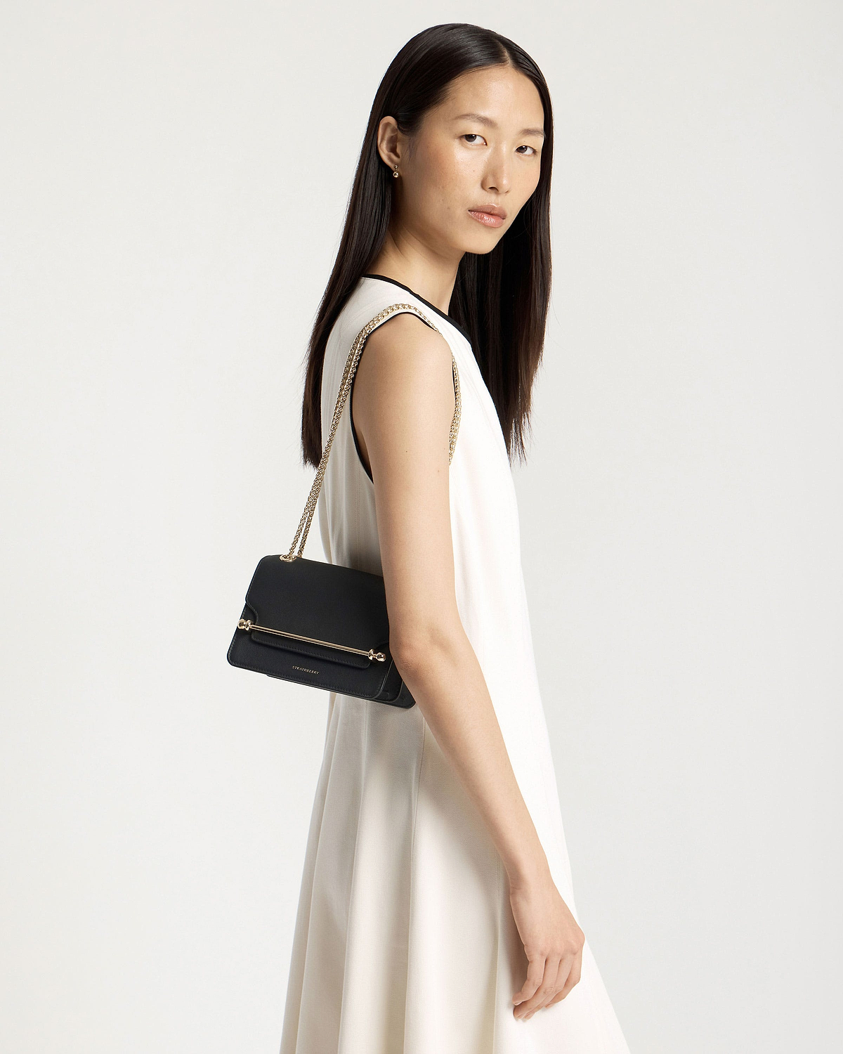 Model wearing white sleeveless dress carrying black Strathberry East/West Mini bag with gold chain strap.