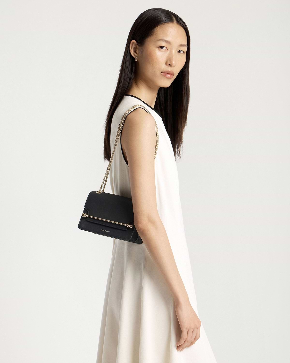 Model wearing white sleeveless dress carrying black Strathberry East/West Mini bag with gold chain strap.