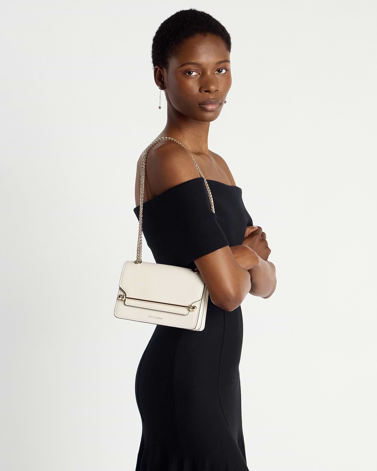 Model wearing black off-shoulder dress with vanilla East/West Mini bag on shoulder.