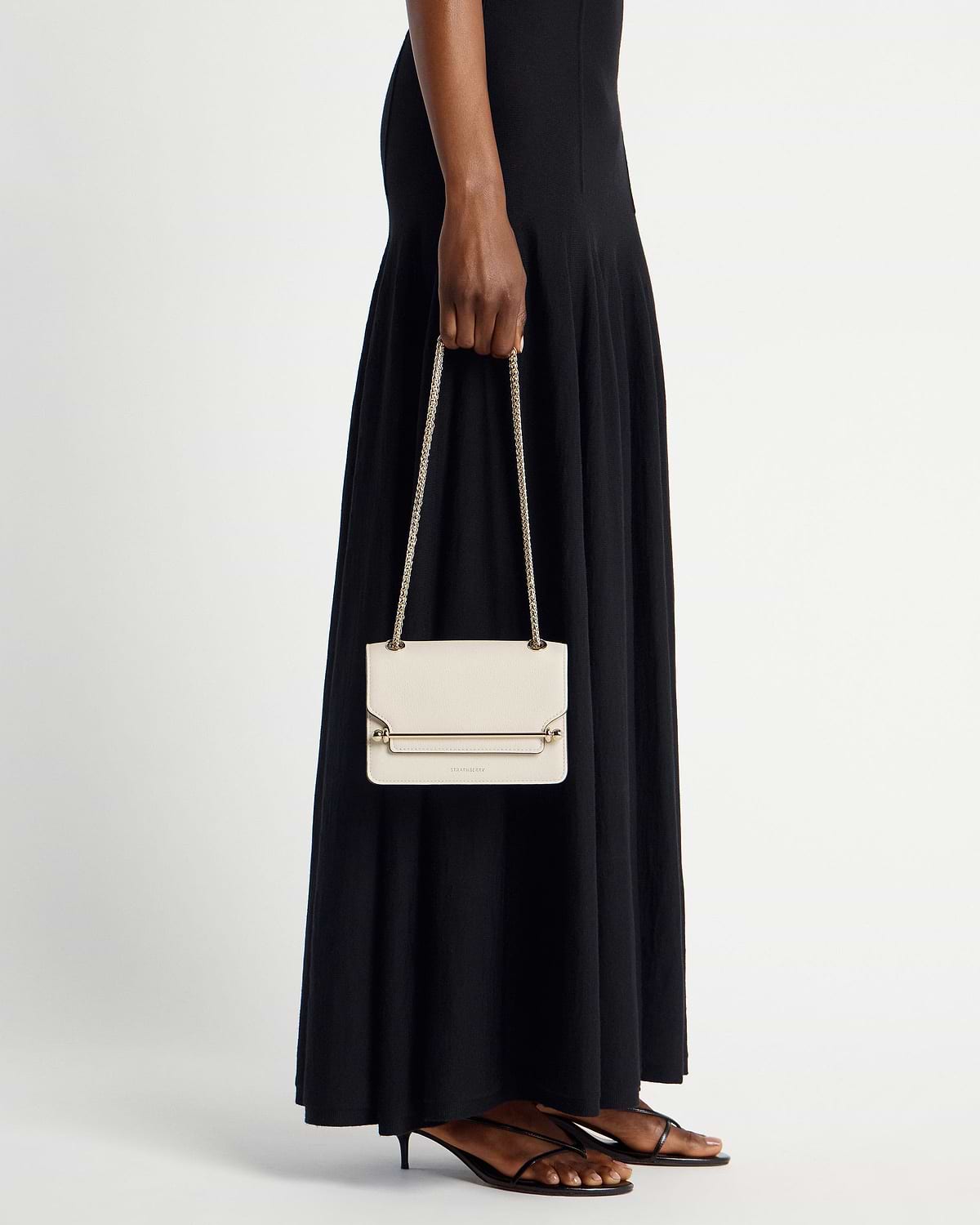 Model holding vanilla East/West Mini bag by Strathberry with chain strap.
