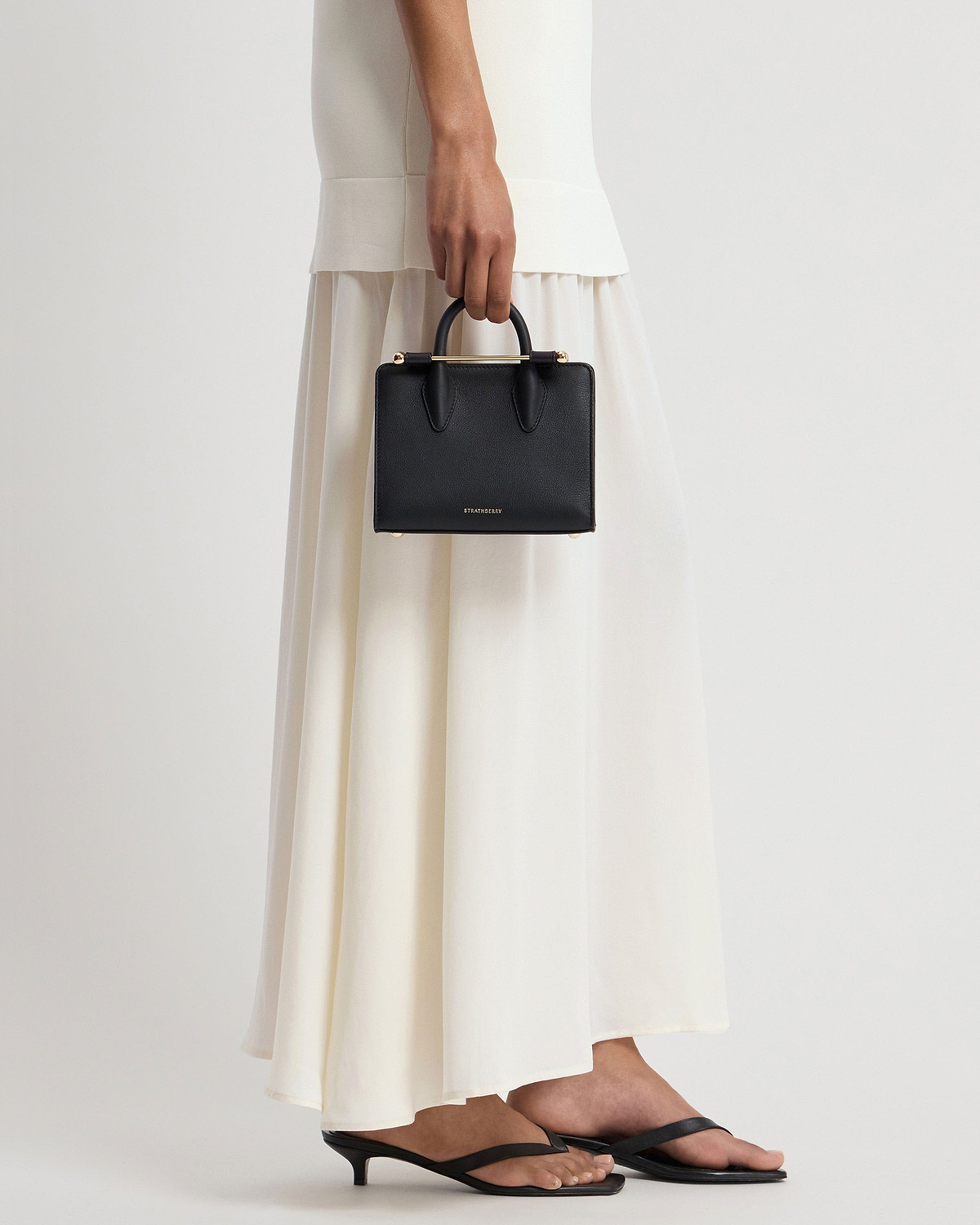 Model holding black Strathberry nano tote with cream-coloured skirt and black sandals.