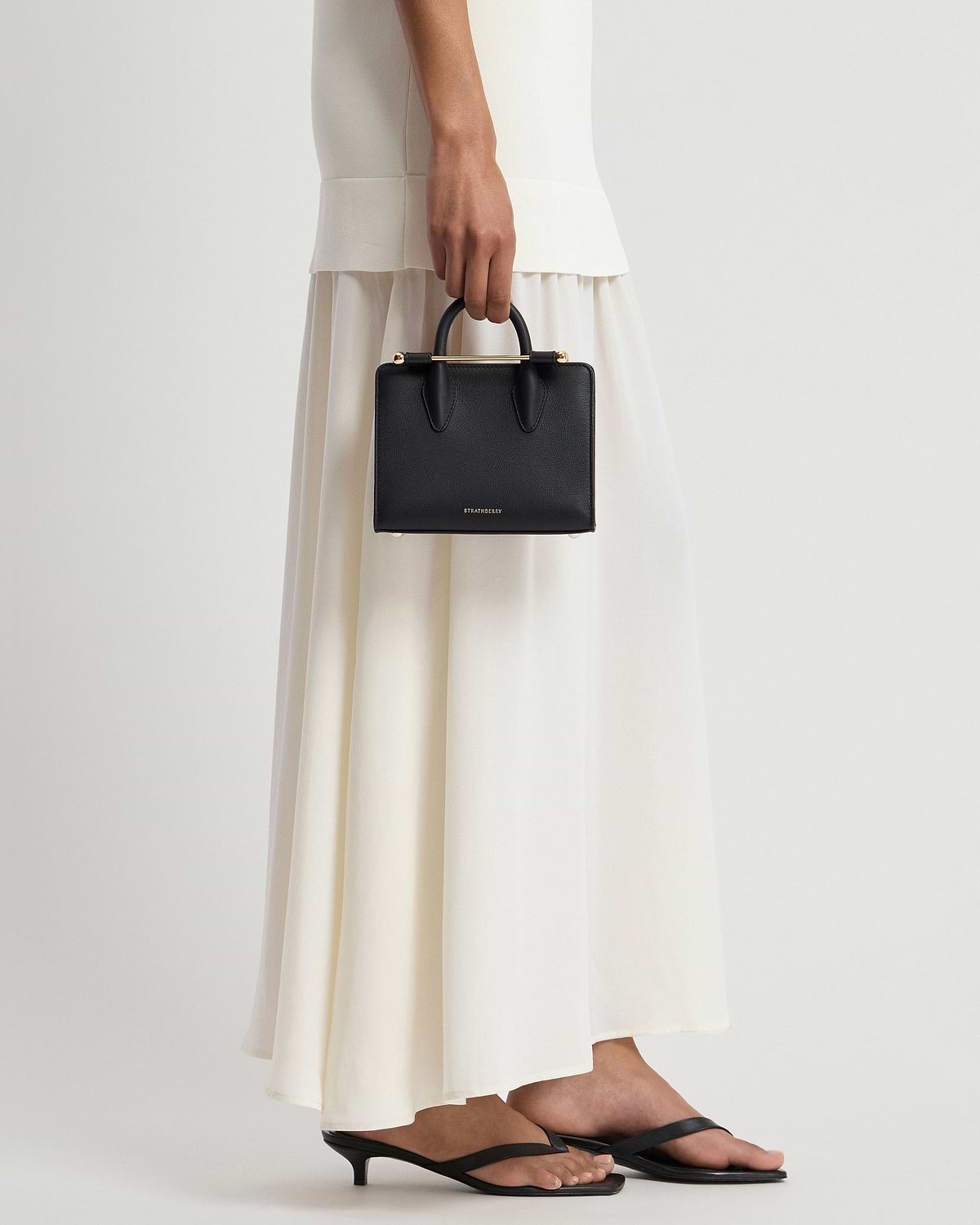 Model holding black Strathberry nano tote with cream-coloured skirt and black sandals.