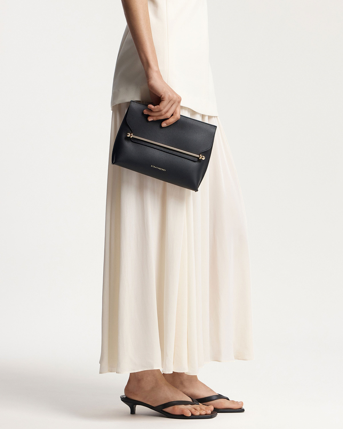 Model waist down holding black Strathberry Stylist clutch with gold bar.