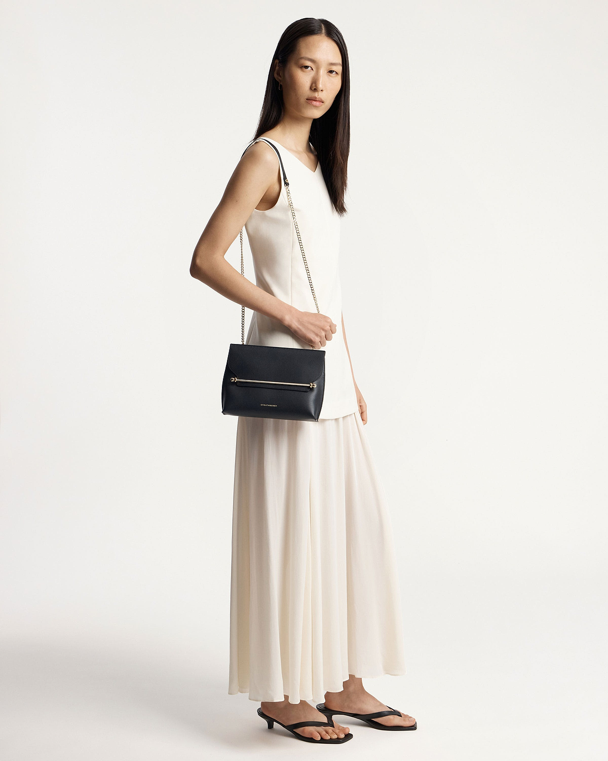 Model wearing a white dress carrying a black Strathberry shoulder bag.