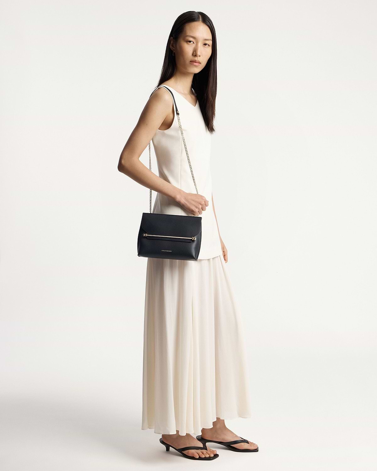 Model wearing a white dress carrying a black Strathberry shoulder bag.