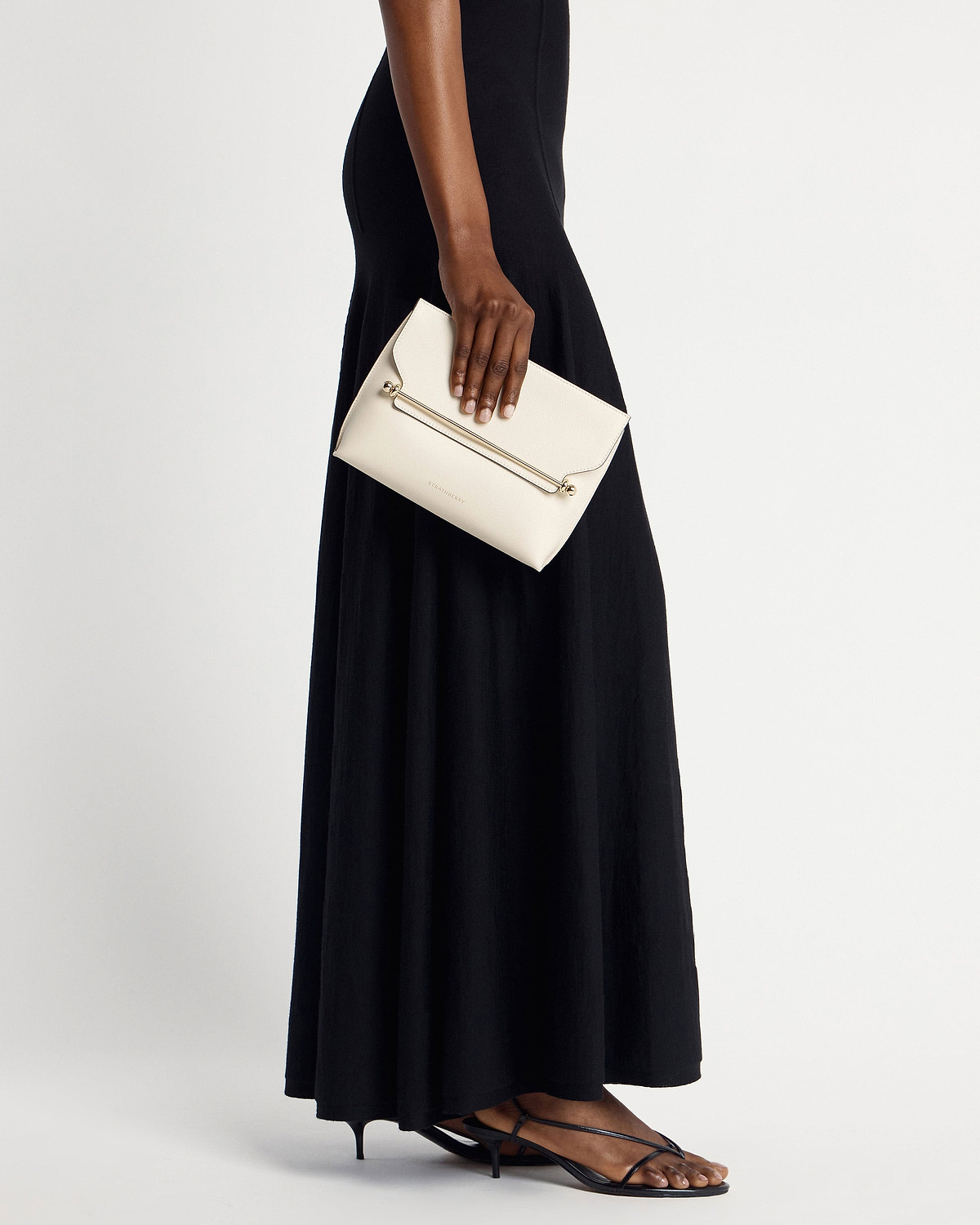 Model's hand holding vanilla Strathberry Stylist clutch with gold bar, black dress visible