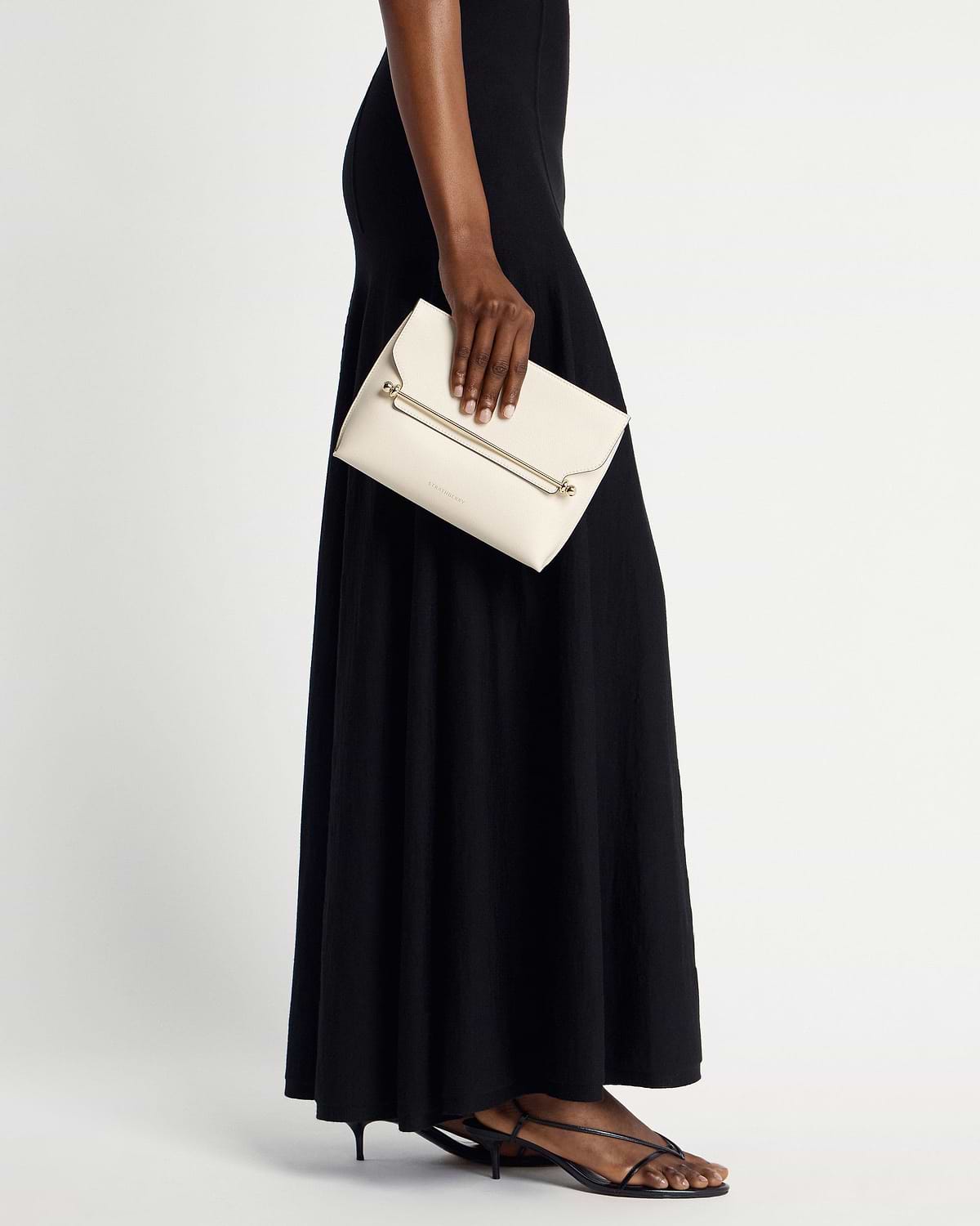Model's hand holding vanilla Strathberry Stylist clutch with gold bar, black dress visible