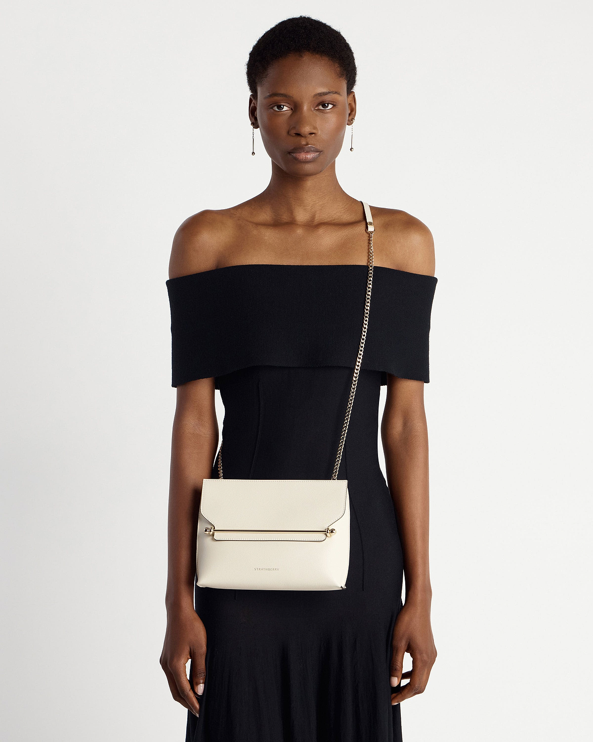 Model in black off-shoulder dress wearing Strathberry Stylist bag in vanilla with gold chain.