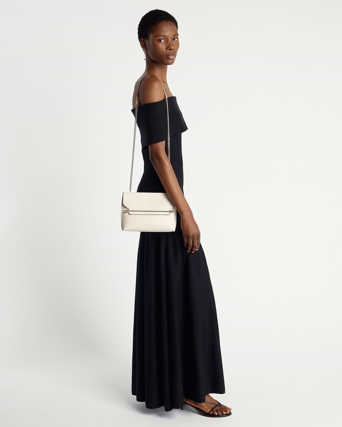 Model wearing black dress carrying vanilla shoulder bag with chain strap.