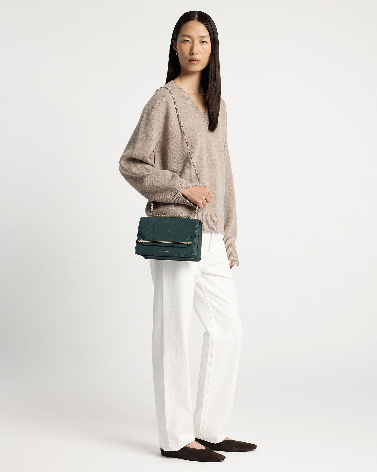 Woman wearing beige jumper and white trousers carrying bottle green east/west bag.