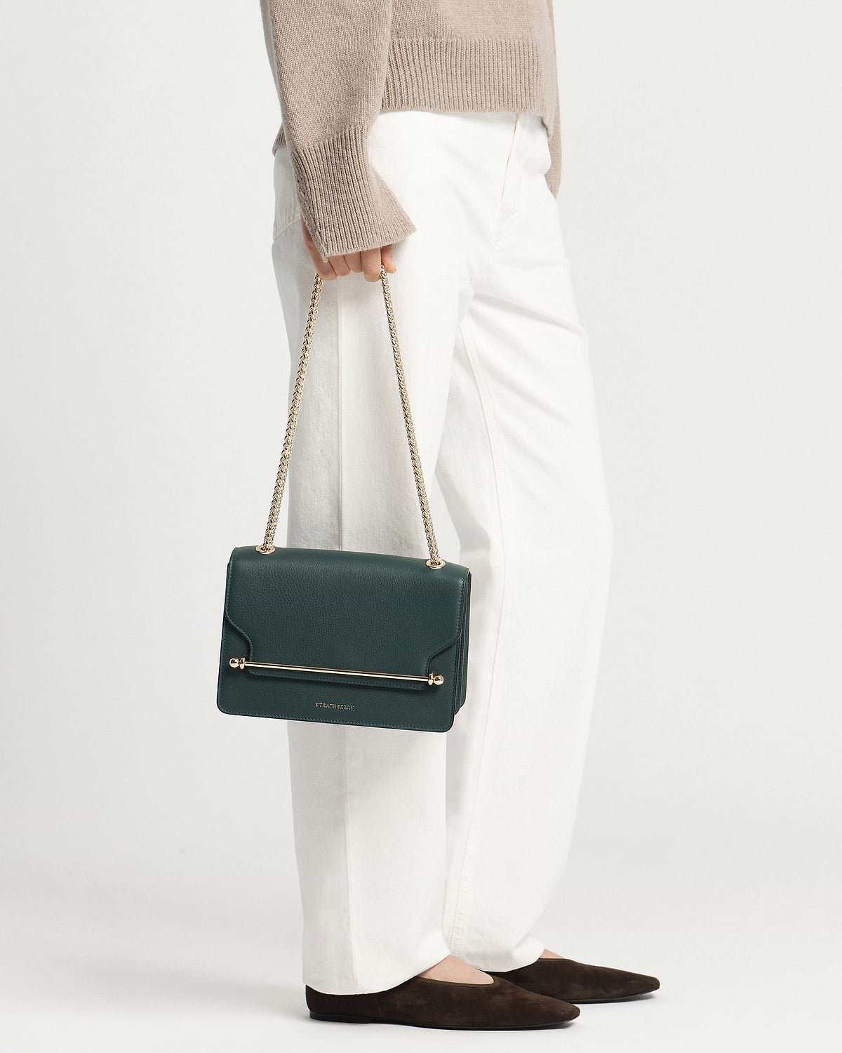 Model holding bottle-green Strathberry East/West bag with gold chain strap.