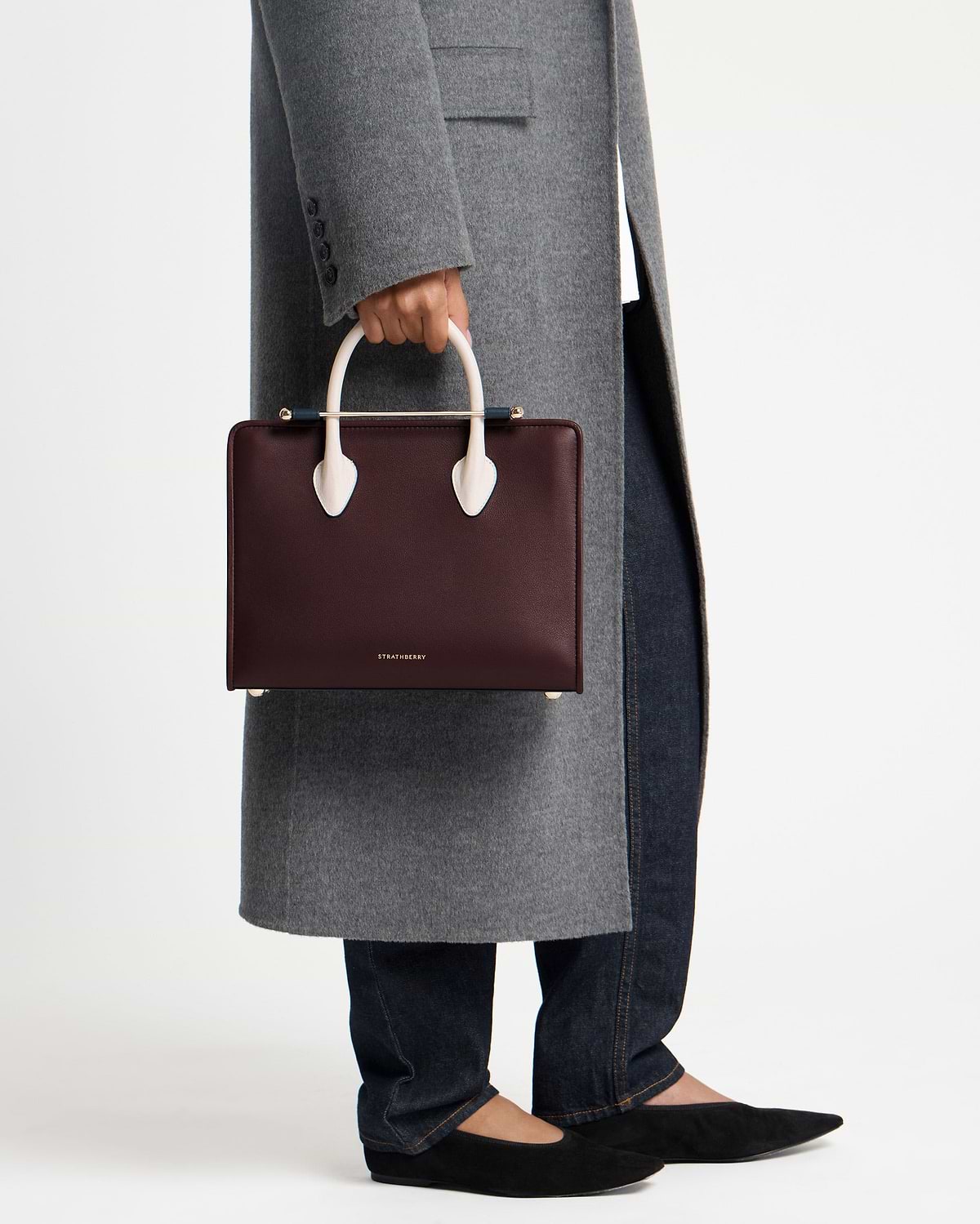 Model cropped below waist holding Strathberry midi tote in burgundy with vanilla and navy handles.