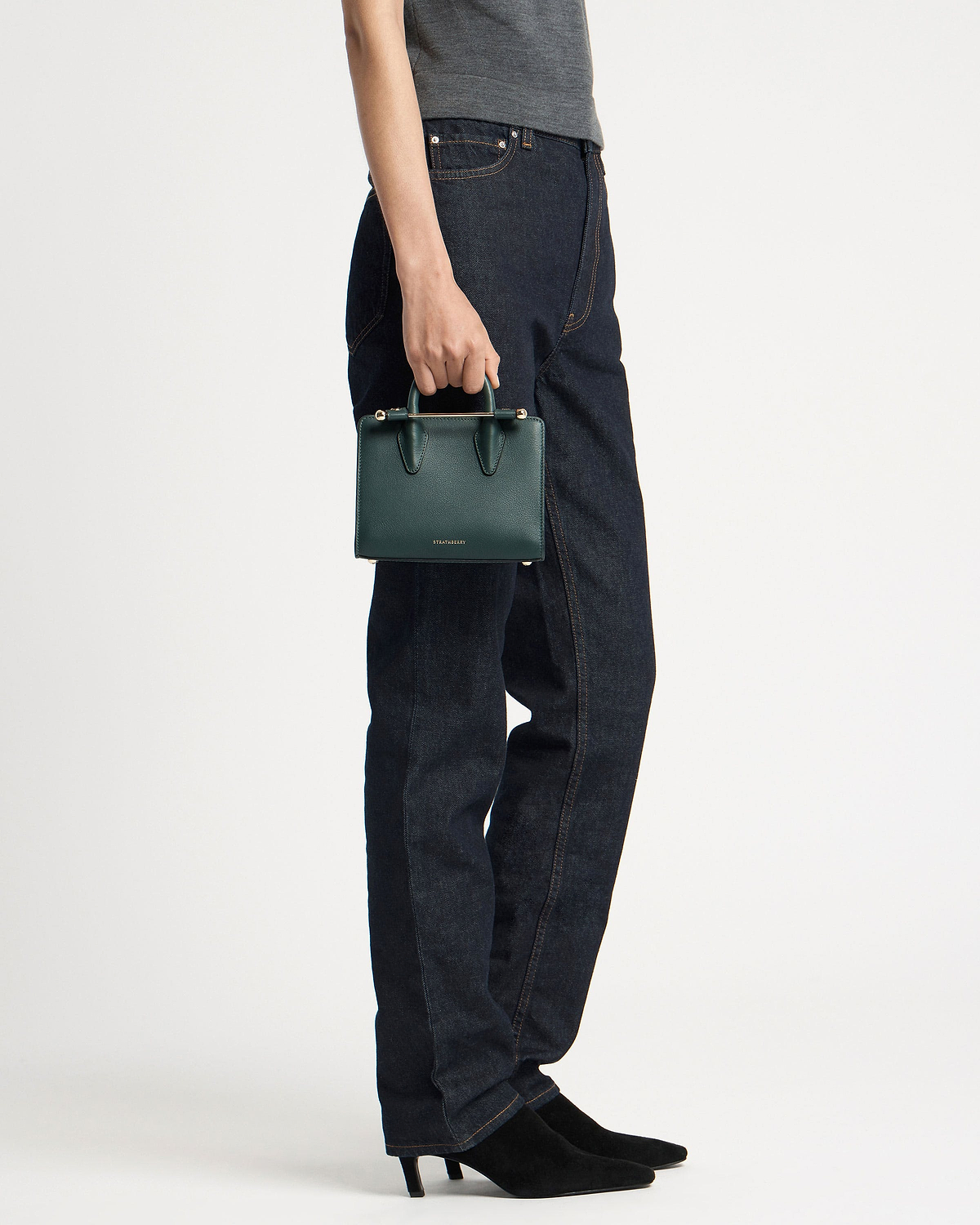Model holding bottle green nano tote by top handle beside dark jeans.