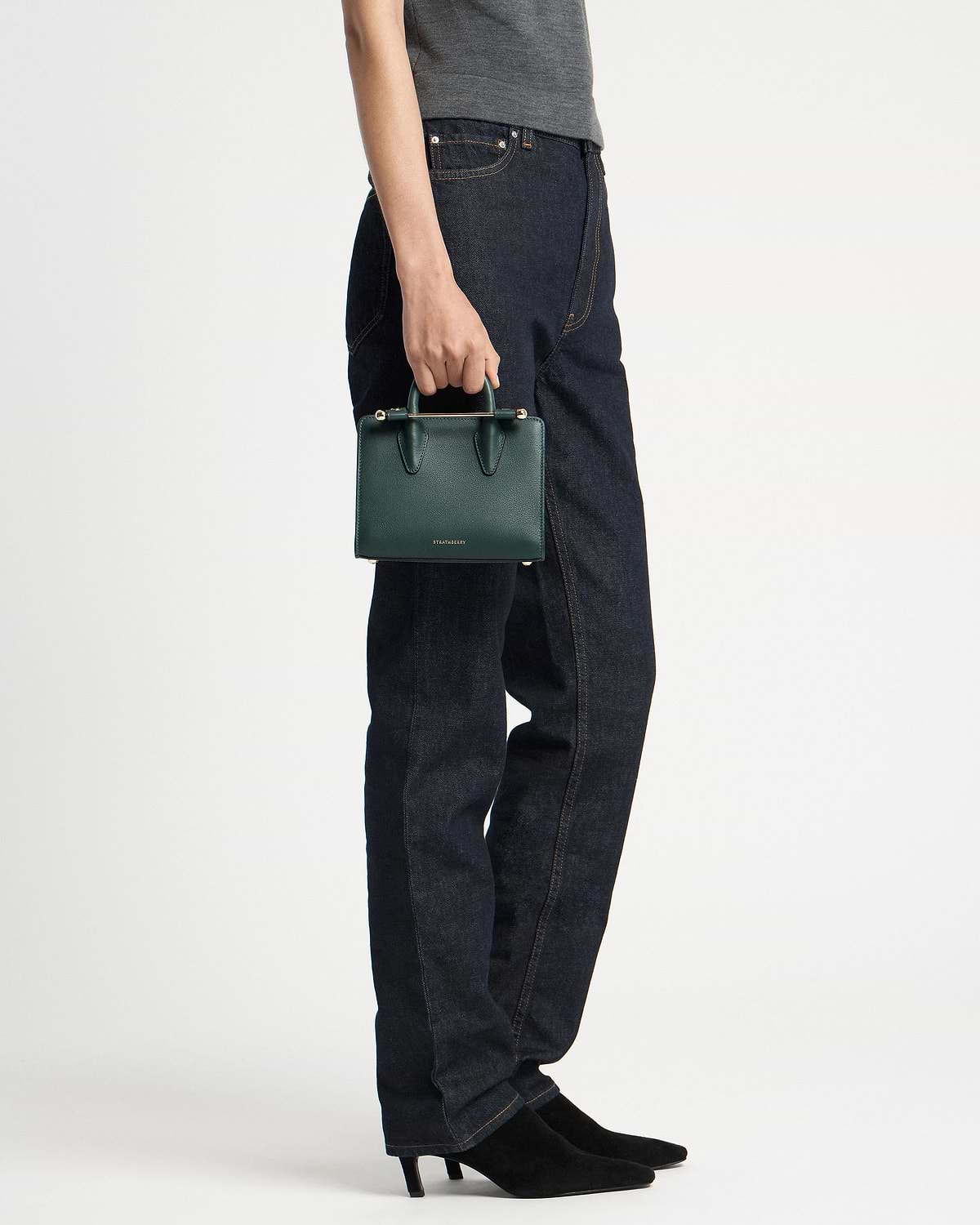 Model holding bottle green nano tote by top handle beside dark jeans.