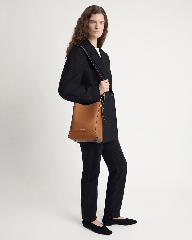 Model standing wearing black blazer and trousers, carrying tan bucket bag with vanilla stitch.
