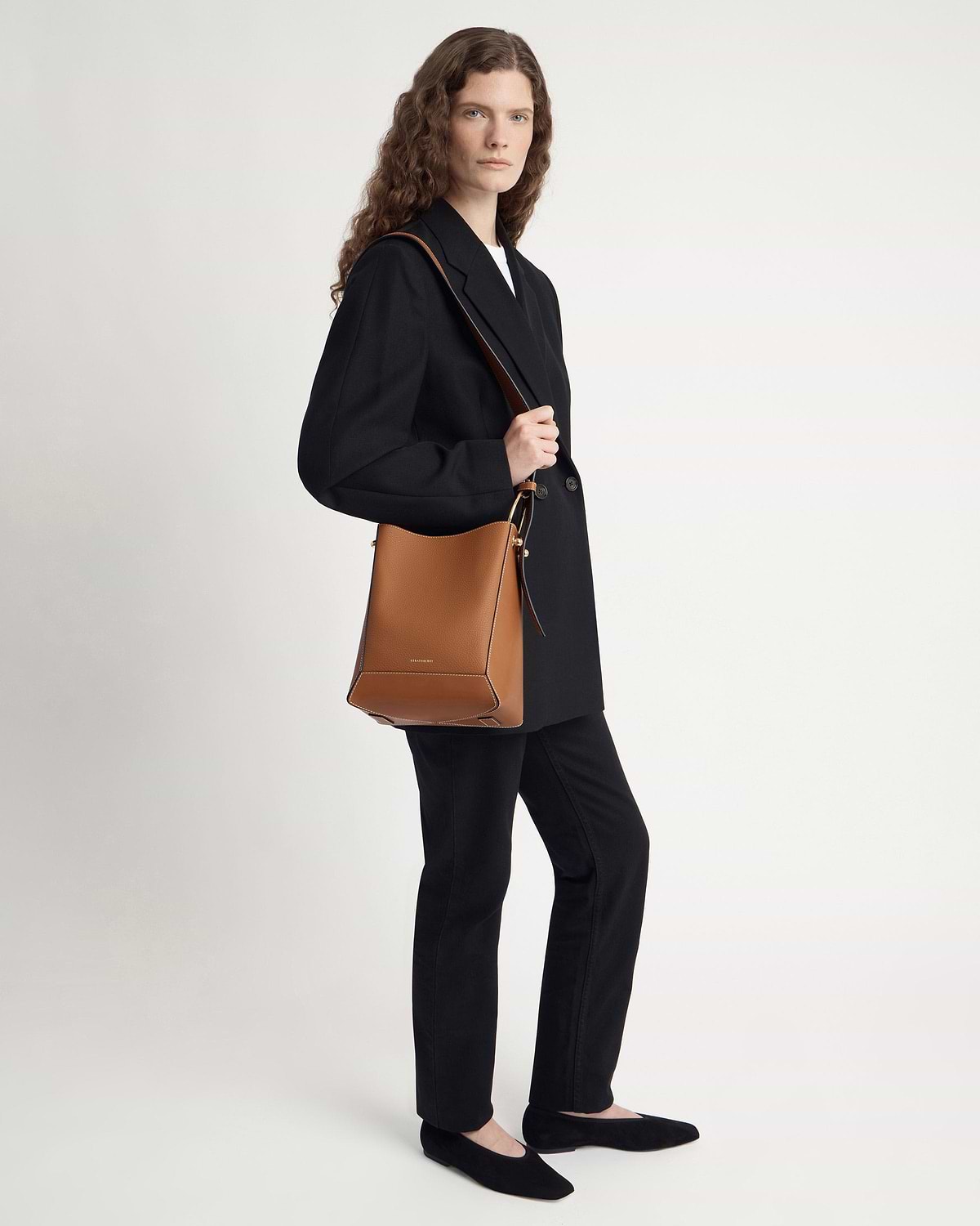 Model standing wearing black blazer and trousers, carrying tan bucket bag with vanilla stitch.