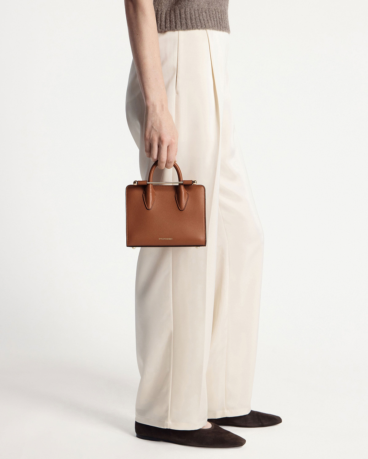 Model holding chestnut Strathberry nano tote by the handles beside cream trousers.