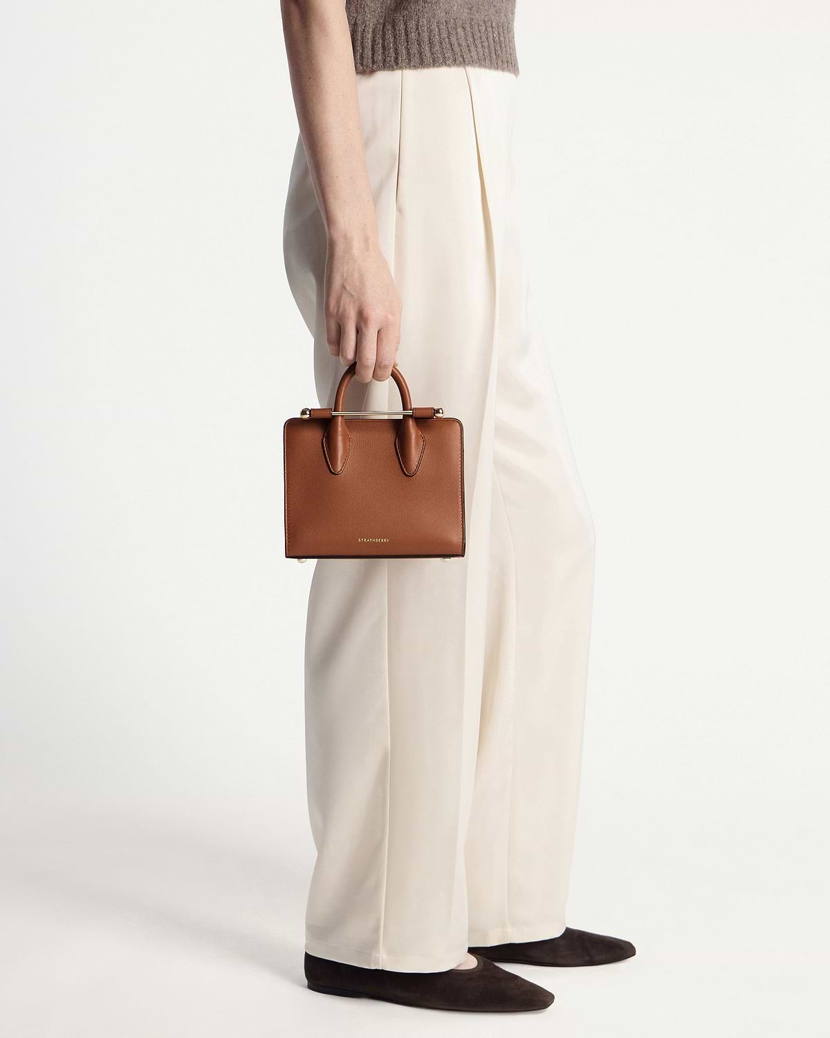 Model holding chestnut Strathberry nano tote by the handles beside cream trousers.
