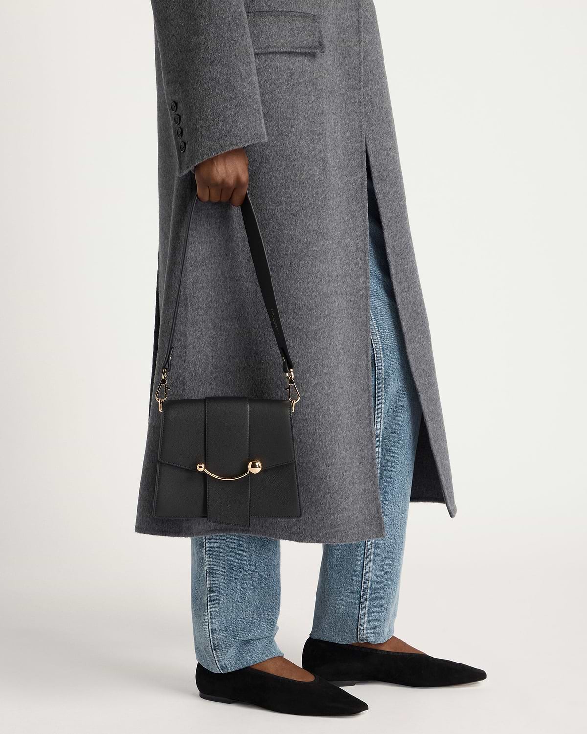 Person holding black box crescent bag with curved gold hardware, grey coat and jeans