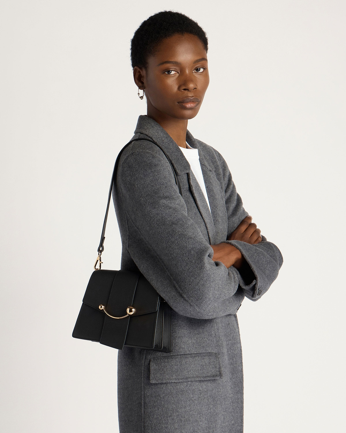 Model wearing grey coat carrying black Box Crescent shoulder bag with gold hardware