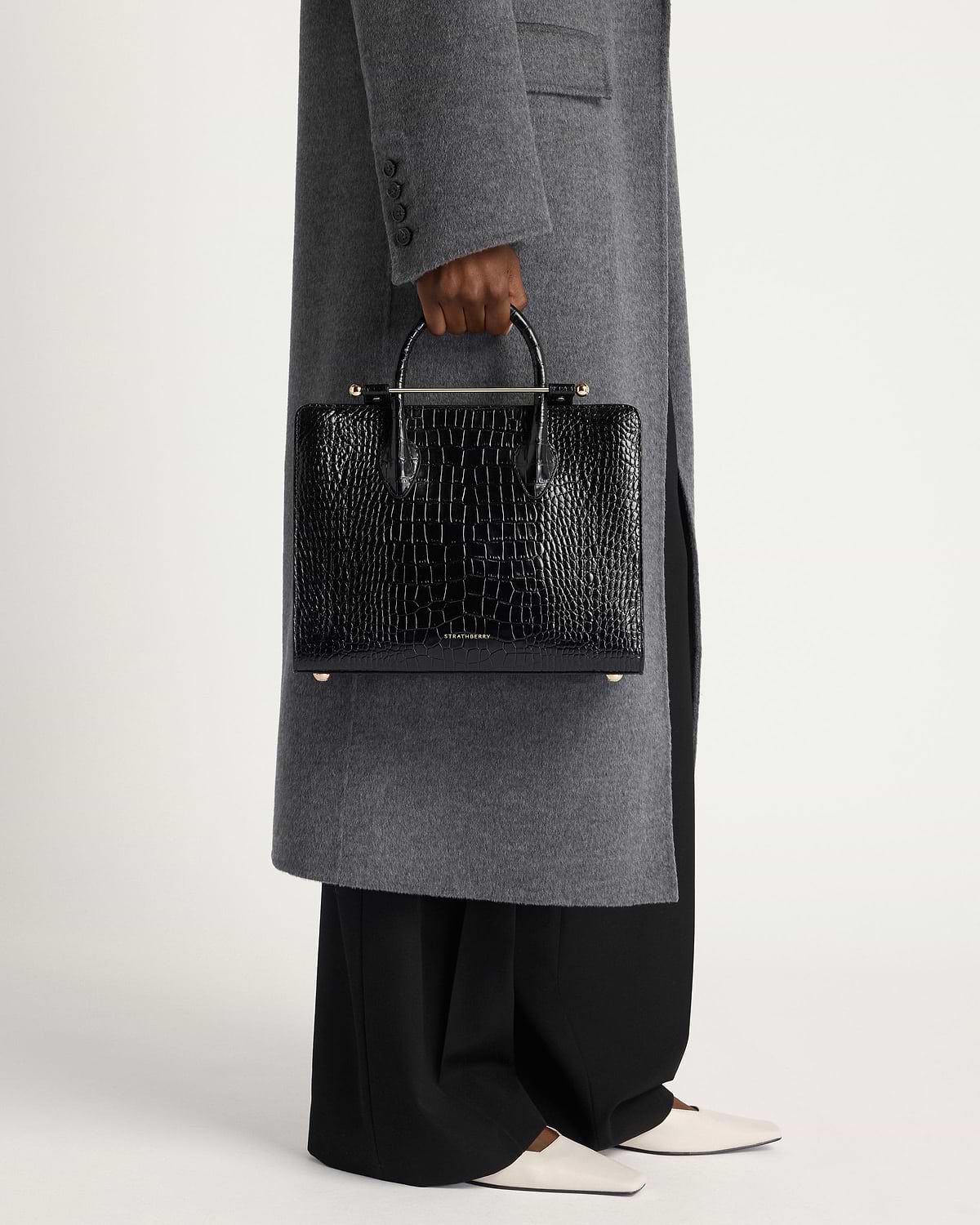 Model holding Strathberry midi tote in black crocodile-embossed leather, worn with grey coat.