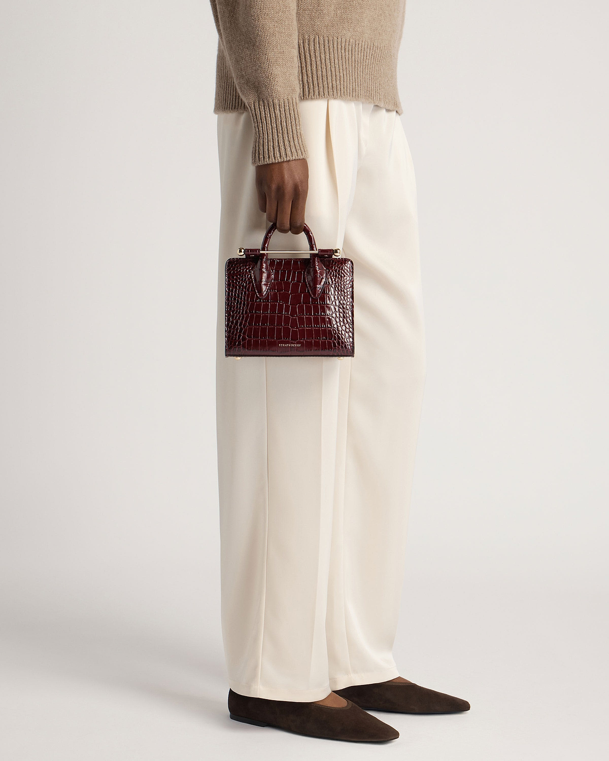 Model holding burgundy croc-effect nano tote by Strathberry at her side.