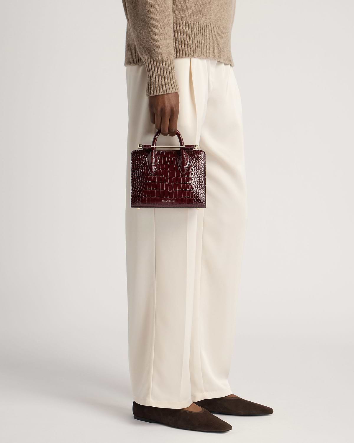Model holding burgundy croc-effect nano tote by Strathberry at her side.