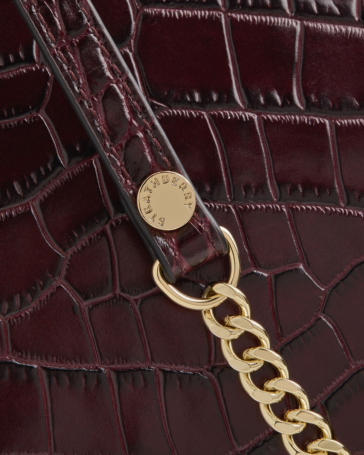 Burgundy croc-embossed leather bag detail with gold chain and Strathberry badge.