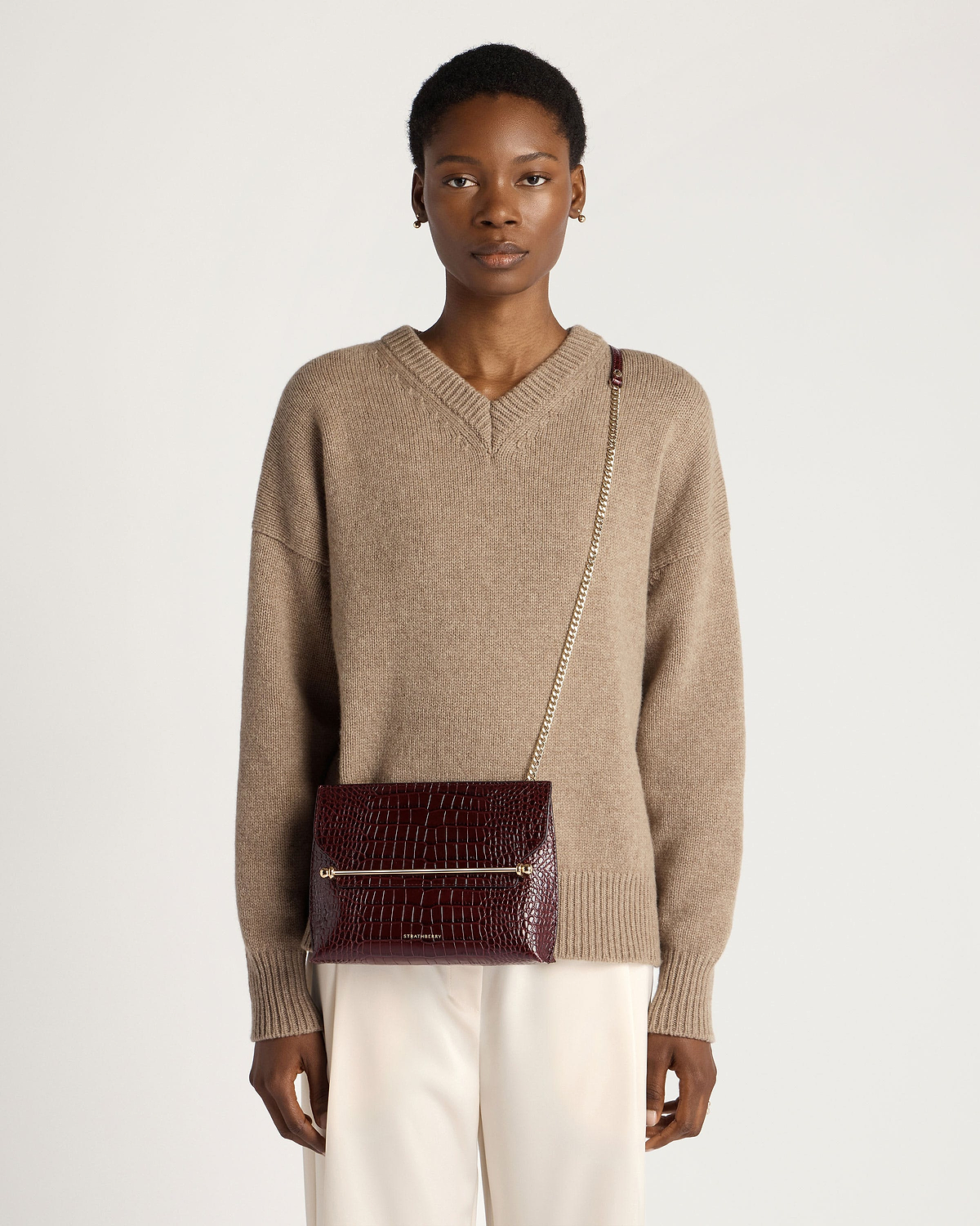 Model wearing Strathberry Stylist croc-effect burgundy crossbody bag with gold chain.
