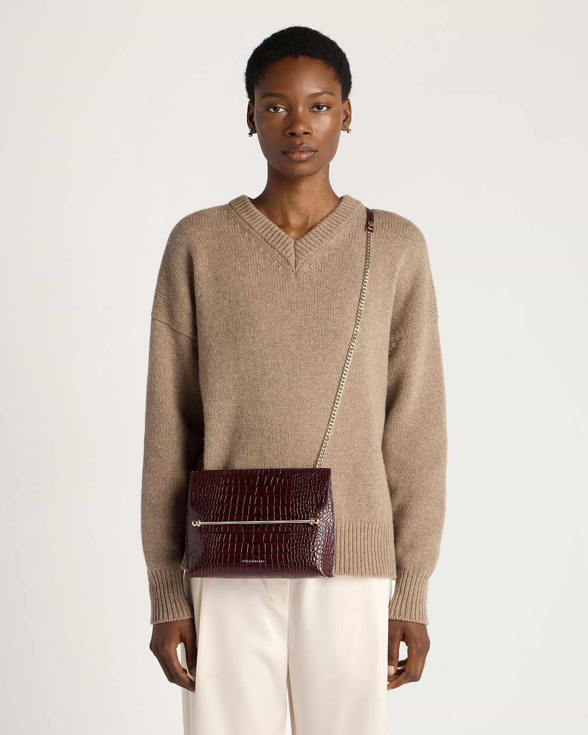 Model wearing Strathberry Stylist croc-effect burgundy crossbody bag with gold chain.