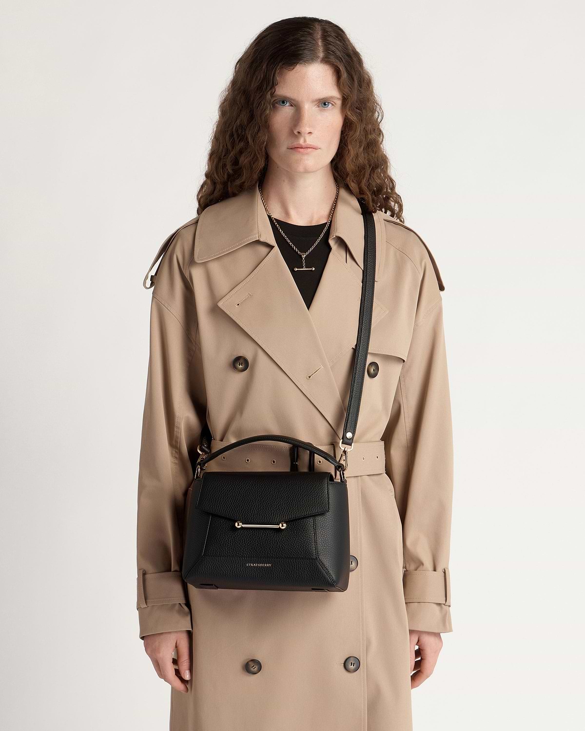 Woman in beige trench coat carrying black Mosaic bag with top handle and shoulder strap.