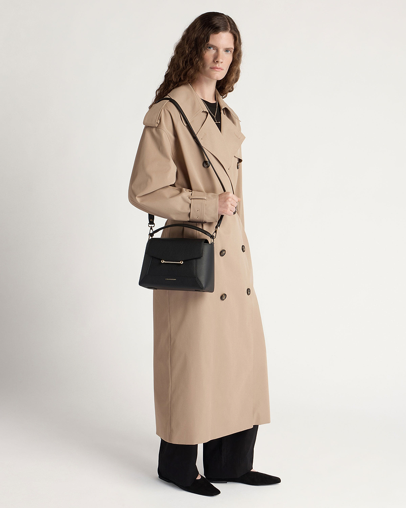 Model in a beige trench coat holding a black mosaic bag with shoulder strap.